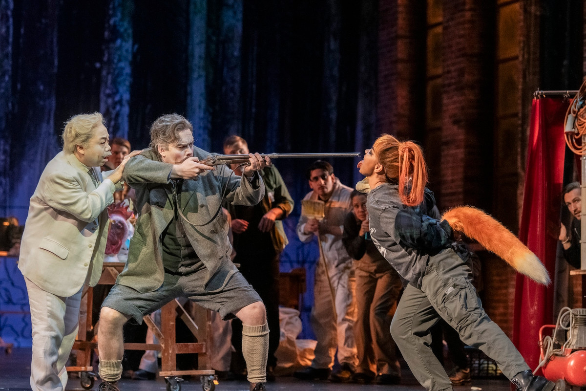 A scene from an opera where a man holds a shotgun against an actor dressed as a fox.