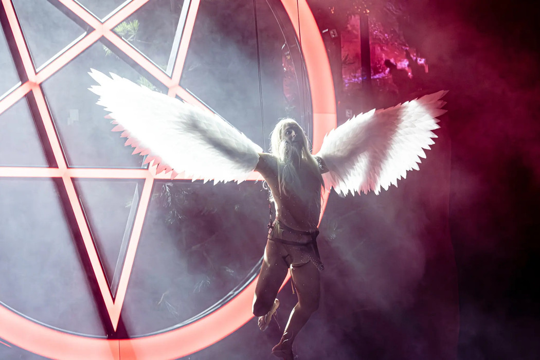 An actor dressed as an angel, hanging from the ceiling in front of a inverted pentagram on a brightly lit stage. 