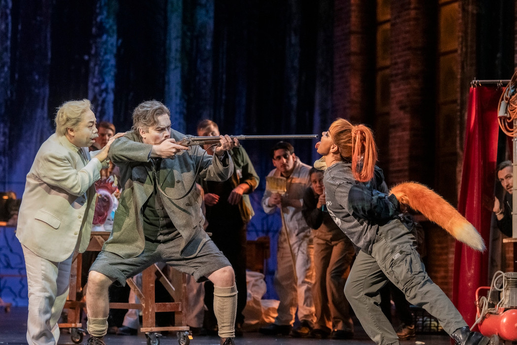 A scene from an opera where a man holds a shotgun against an actor dressed as a fox. 
