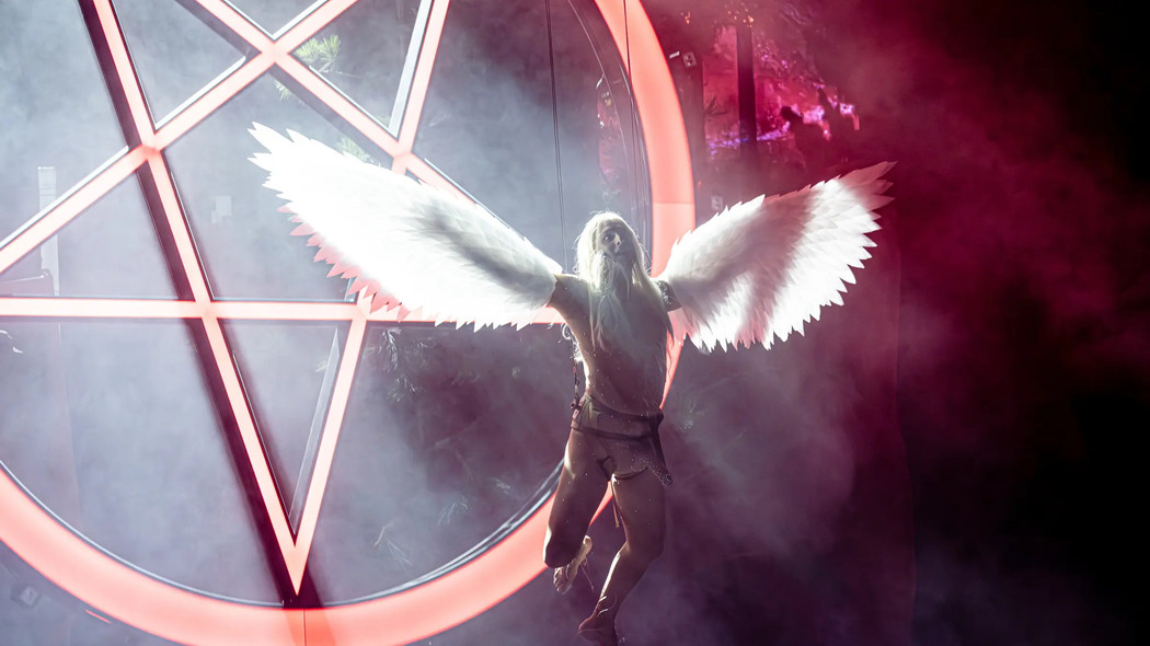 An actor dressed as an angel, hanging from the ceiling in front of a inverted pentagram on a brightly lit stage. 